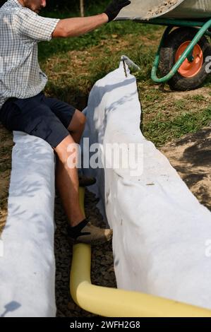 Un lavoratore si trova in una fossa di drenaggio con geotessile bianco. Piedi dell'artigiano sulle macerie di granito da vicino. Foto Stock