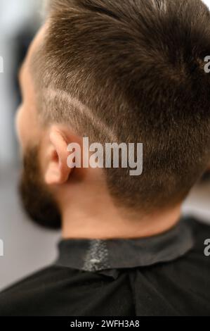 Un giovane con la barba in un mantello nero siede su una sedia in un barbiere. Il cliente sta aspettando un taglio di capelli e la rasatura della barba. Foto Stock