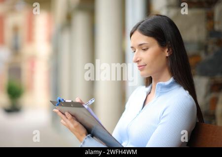 Donna seria che compila il modulo seduta su una panchina per strada Foto Stock