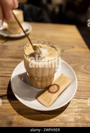 Gustando un cappuccino appena fatto, mescolando il latte con la schiuma in una tazza di vetro trasparente su un tavolo rustico in legno. Foto Stock