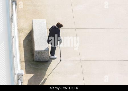 Donna anziana con una stampella vicino a una panchina in una strada che guarda il terreno. Copia spazio. Vista ad alto angolo Foto Stock