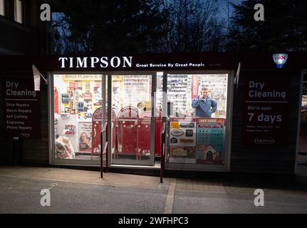 Torquay. Negozio di scarpe Timpson, riparazione orologi e taglio chiavi con il ciabattino all'interno al telefono. Fotografia notturna con negozio illuminato al buio. Foto Stock