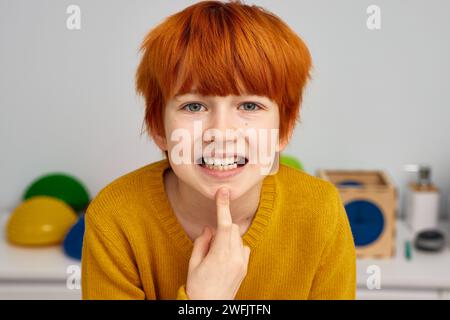 Ritratto di un ragazzo dai capelli rossi che pronuncia lettere S nell'ufficio del logoterapista. Terapia del linguaggio e del linguaggio per bambini Foto Stock