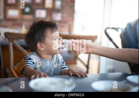 Un simpatico bambino multirazziale seduto al tavolo di un ristorante su una sedia alta, mentre sua madre lo nutre con il cucchiaio durante il pranzo Foto Stock