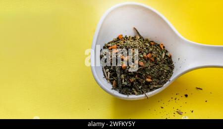 Cucchiaio bianco in ceramica con foglie di tè secche e varie aggiunte di bacche su sfondo giallo Foto Stock