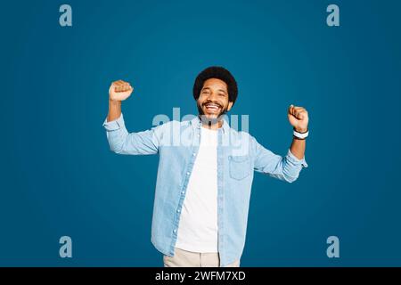 Giovane con un sorriso allegro allegra entrambi i pugni in un gesto trionfale, mostrando un momento di successo o di celebrazione su uno sfondo blu. Lo scatto enfatizza le sensazioni di soddisfazione e gioia Foto Stock