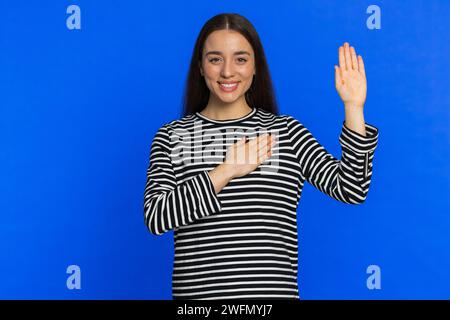 Giuro di essere onesto. Donna sorridente, sincera e responsabile, che si alza la mano per giurare, promettendo di essere onesta e di dire la verità, tenendo la mano sul petto. Ragazza isolata sullo sfondo blu dello studio. Stili di vita Foto Stock