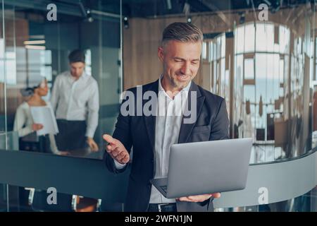 L'affascinante uomo d'affari che parla tramite videochiamata con il cliente utilizza un computer portatile mentre si trova in ufficio Foto Stock