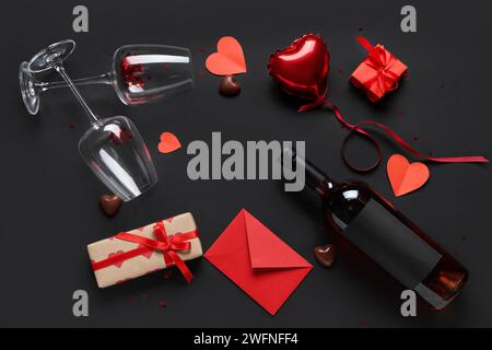 Cornice realizzata con cuori di carta con bottiglia di vino, bicchieri, confezioni regalo, busta, palloncino ad aria compressa e caramelle di cioccolato su sfondo nero. San Valentino Foto Stock