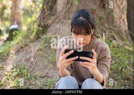 La giovane donna asiatica si concentra su un gioco sul suo smartphone mentre si siede sotto un albero in un parco. concetti relativi alle persone e alla tecnologia wireless Foto Stock