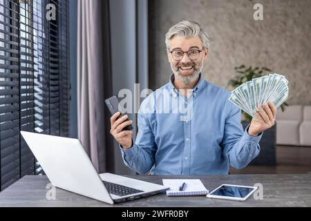 Uomo d'affari che ama le bollette del dollaro e lo smartphone mentre è seduto in un ufficio domestico con un computer portatile. Uomo maturo gioioso che si eccita per un reddito massiccio dopo una giornata lavorativa produttiva. Foto Stock