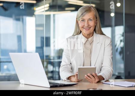 Donna bionda anziana che utilizza un tablet digitale mentre si siede a un tavolo con un computer portatile sullo sfondo dello spazio di lavoro. Donna d'affari sicura di sé che utilizza gadget moderni aggiuntivi per aumentare la produttività. Foto Stock