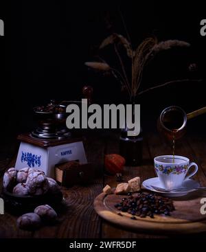 Composizione del caffè. Il caffè viene versato da una tazza turca in una tazza, un macinacaffè, caffè macinato e chicchi di caffè, biscotti croccanti al cioccolato e secchi Foto Stock