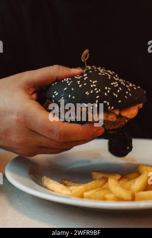 Mano maschile che tiene un hamburger di manzo con un panino nero in un caffè. Delizioso pranzo americano Foto Stock