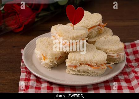 Panini a forma di cuore fatti di insalata seduti su un piatto bianco su un panno a scacchi rosso e bianco. Sullo sfondo c'è un bouquet di rose. saint, va Foto Stock