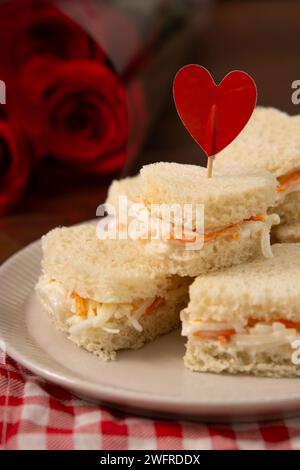 Panini a forma di cuore fatti di insalata seduti su un piatto bianco su un panno a scacchi rosso e bianco. Sullo sfondo c'è un bouquet di rose. saint, va Foto Stock