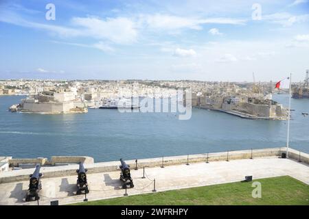 Una vista panoramica della storica città fortificata di la Valletta e del vivace porto sotto un cielo azzurro, con i cannoni in primo piano come un cenno ai suoi piani Foto Stock