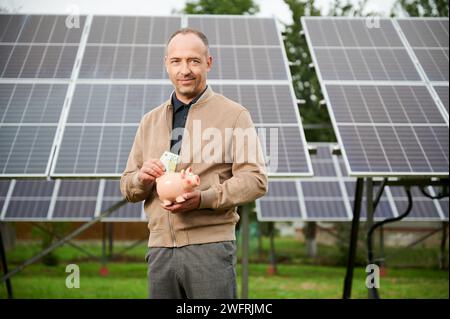 Uomo di fiducia che mette i soldi guadagnati alla banca del piggy. Il profitto di risparmio dell'adulto maschio ha ottenuto investendo nell'energia verde. Uomo in abiti casual tenendo in mano la scatola del denaro e mettere euro in esso. Foto Stock