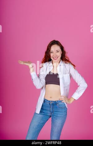 Felice donna rossa con una camicia bianca che si presenta con la mano, in piedi con sicurezza su uno sfondo rosa. Foto Stock