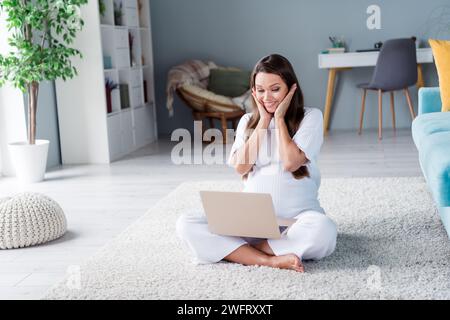 Foto di una ragazza incinta eccitata felice allegra e positiva seduta sul pavimento a guardare video in un confortevole appartamento la mattina al chiuso Foto Stock