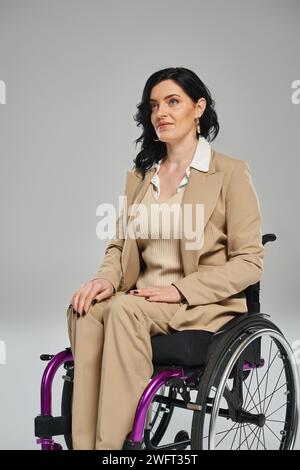 donna bruna sicura con disabilità in abito pastello seduta in sedia a rotelle e guardando lontano Foto Stock