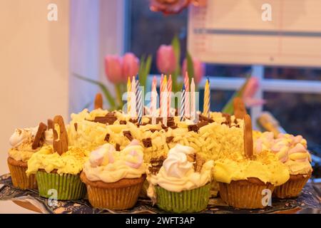 Una torta di compleanno al cioccolato bianco con candele spente circondata da cupcake fatti in casa coperti da decorazioni individuali e glassa di burro Foto Stock
