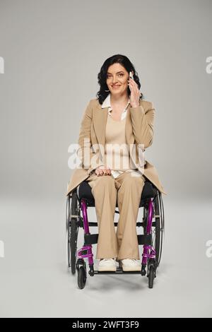 donna allegra e attraente con disabilità in sedia a rotelle che parla al telefono e guarda la fotocamera Foto Stock