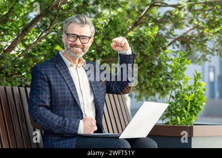 Un dirigente uomo maturo sicuro di sé in una tuta aziendale celebra il successo con la pompa a pugno, utilizzando un notebook all'aperto. Foto Stock