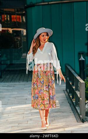 Giovane donna alla moda con cappello, lunghi capelli biondi, blusa bianca e fiorita, lunga gonna a pieghe. Foto Stock