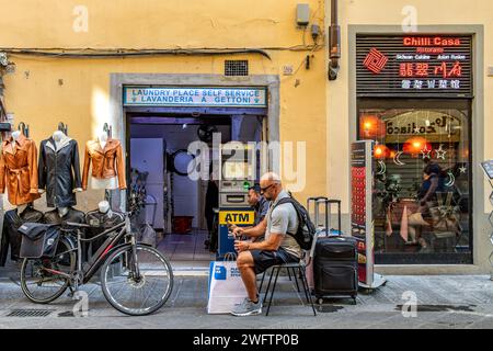 Una donna che usa una lavatrice, lavanderia a gettoni, con due uomini seduti fuori ad aspettare in via Faenza, una strada a Firenze, Italia Foto Stock