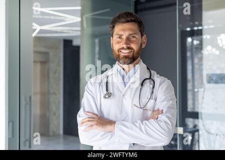 Ritratto ravvicinato di un giovane medico maschio sorridente, assistente, studente in piedi nello studio dell'ospedale e guardando con sicurezza la telecamera con le braccia incrociate sul petto. Foto Stock
