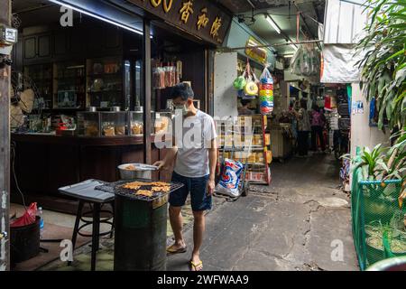 Un uomo ha visto di fronte al suo negozio cinese grigliare delle fette di maiale su un barbecue, in uno dei vicoli del mercato di Khlong Toei, in Rama IV Road, nel quartiere di Khlong Toei. Il mercato di Khlong Toei è il più grande mercato fresco di Bangkok. È diventato un punto focale dell'attività economica, con una vasta gamma di venditori che vendono frutta fresca, verdura, pesce, carne e altri beni essenziali, che fungono anche da luogo di incontro per le comunità locali. Foto Stock