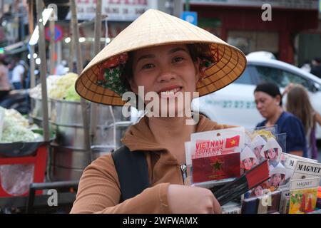 Donna sorridente in cappello conico che vende souvenir per strada fuori da un mercato affollato a ho chi Minh, Vietnam Foto Stock