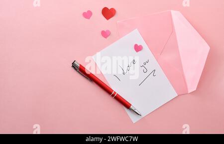 Nota scritta i Love You su carta bianca su sfondo rosa con penna su busta rosa e ritagli di carta a forma di cuore. Vista dall'alto. Foto Stock