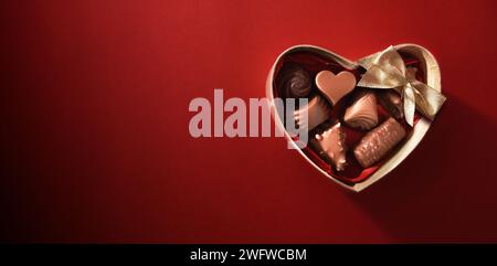 Scatola marrone a forma di cuore piena di cioccolatini assortiti con fiocco dorato su sfondo rosso sfumato. Vista dall'alto. Foto Stock