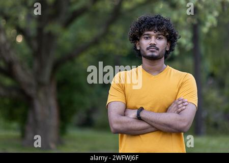 Ritratto di un atleta indiano serio e sicuro che si trova all'aperto in un parco in abiti casual, guardando la fotocamera con le braccia incrociate sul petto. Foto Stock