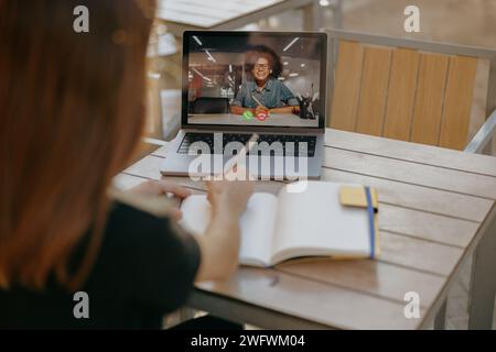 Una manager di sesso femminile parla con il cliente tramite videochiamata e prende appunti mentre è seduto nella terrazza del bar Foto Stock