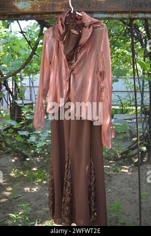 Vita ordinaria, stile, moda, bellissimo abito da donna marrone con giacca organza marrone rossa appesa su un appendiabiti bianco nel cortile della casa. Foto Stock