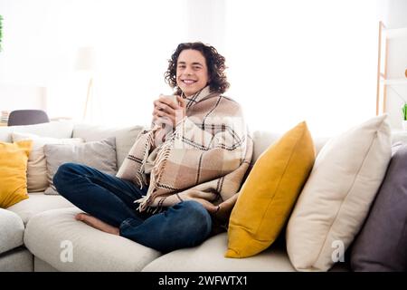 Foto di un ragazzo eccentrico e allegro che gira la coperta a quadri congelando gustando una gustosa bevanda calda all'interno della casa Foto Stock