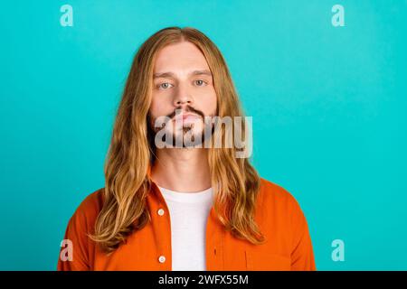 Il ritratto di un giovane ragazzo con lunghi capelli biondi dritti che indossa una camicia arancione sembra davvero serio isolato su sfondo color acquamarina Foto Stock