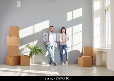 Coppie di famiglie felici in abiti informali sono in piedi insieme vicino alle pareti sotto i raggi di luce solare tra le scatole di cartone. Coppia sorridente sposata compra o affitta Foto Stock