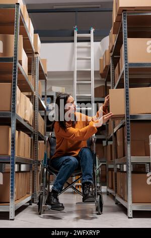 Storehouse asian commissionatore su sedia a rotelle che prende il pacco dal rack al confezionamento del prodotto prima della spedizione. Giovane donna che gestisce il magazzino di vendita al dettaglio che sceglie la scatola sullo scaffale Foto Stock