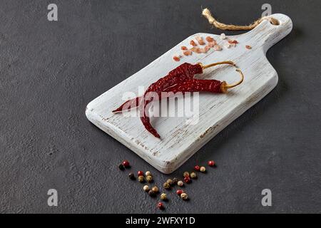 due peperoni rossi caldi, grani di pepe sparsi e sale rosa su un tagliere di legno bianco su sfondo di pietra nera Foto Stock