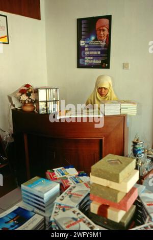 Una donna lavora dietro la cassa, di fronte a un muro dove una foto del fondatore Tahiid Daarut, il leader religioso islamico indonesiano KH Abdullah Gymnastiar (popolarmente noto come AA Gym), è collocata in una libreria vicino alla moschea Daarut Tauhiid a Gegerkalong, Bandung, Giava Occidentale, Indonesia. Fondato da KH Abdullah Gymnastiar, Daarut Tauhiid collegio e fondazione, insieme alla loro gestione di filiali, Manajemen Qolbu (mq), sono riusciti a intensificare il turismo religioso e le attività economiche nell'area di Gegerkalong nella città di Bandung. Foto Stock