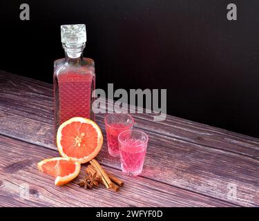 Liquore di pompelmo con spezie su un tavolo di legno, forte alcol fatto in casa in bottiglia e due bicchieri da shot con cannella, anice e pezzi di cit maturo Foto Stock