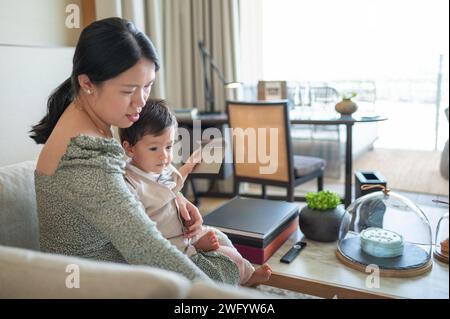 Carino bambino multirazziale di un anno che festeggia il suo primo compleanno con sua madre a casa. È vestito con un abito di lino estivo beige con una brigh Foto Stock