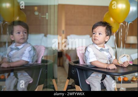 Un bambino multirazziale di un anno che festeggia il suo primo compleanno con palloncini seduti in una sedia da pranzo a casa. È vestito in beige Foto Stock