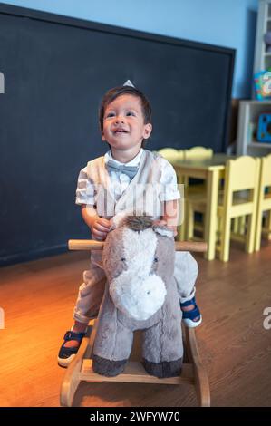 Carino bambino multirazziale di un anno che festeggia il suo primo compleanno all'asilo. Sta cavalcando un cavallo di legno giocattolo e sorride dolcemente Foto Stock