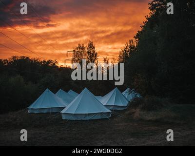 Un gruppo di tende bianche allestite in un campo con un tramonto rosso fuoco sullo sfondo. Foto Stock