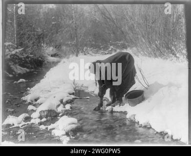 Una giornata invernale ad Apsaroke, c1908. Donna Apsaroke in piedi nella neve che raccoglie l'acqua da un ruscello con una lattina, secchio accanto a lei. Foto Stock